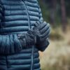 Torso of a person in a dark blue quilted puffer jacket with raindrops, hands clasped in grey insulated gloves against a blurred rainy woodland background.
