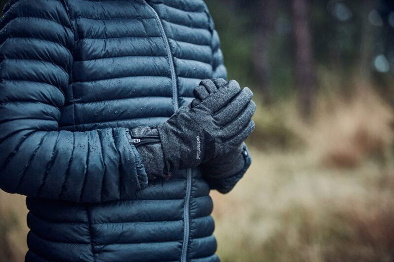 Torso of a person in a dark blue quilted puffer jacket with raindrops, hands clasped in grey insulated gloves against a blurred rainy woodland background.