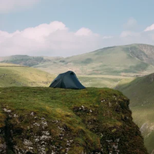 Dark green dome tent pitched near the edge of a grassy cliff, overlooking rolling green hills and a wide valley beneath a pale blue sky with scattered clouds.