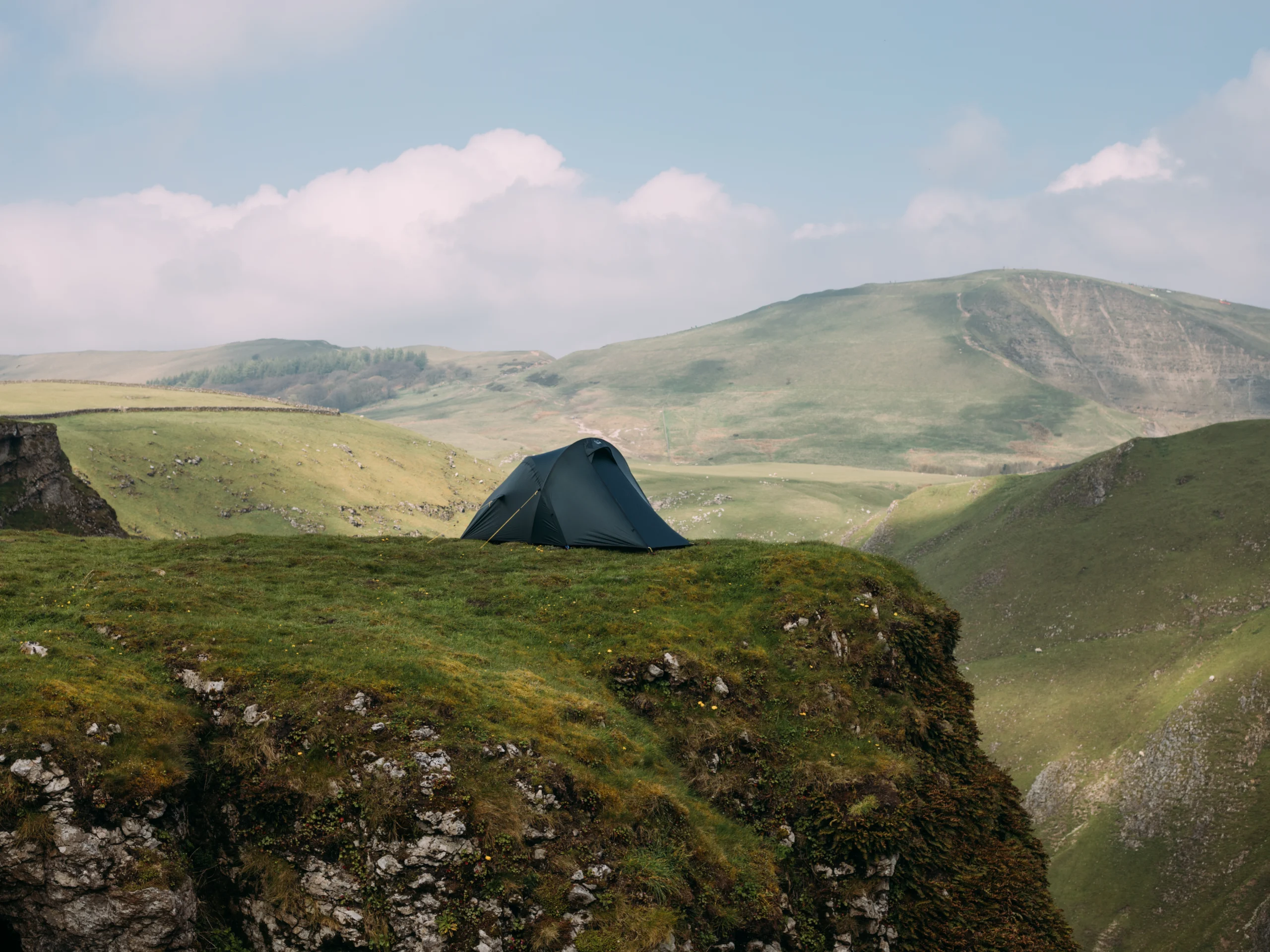 Dark green dome tent pitched near the edge of a grassy cliff, overlooking rolling green hills and a wide valley beneath a pale blue sky with scattered clouds.