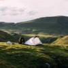 A camper kneeling to pitch a small white tent on a grassy ridge with rolling green hills and a cloudy sky in the background.