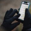 Hands in black gloves holding a smartphone with a green case displaying a detailed topographic map, set against a blurred outdoor background.