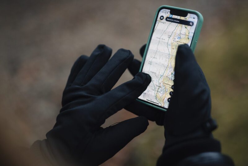 Hands in black gloves holding a smartphone with a green case displaying a detailed topographic map, set against a blurred outdoor background.