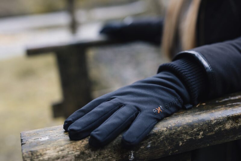 Close-up of a black gloved hand resting on a weathered wooden bench, showing padded fabric, a small orange logo on the glove and a blurred outdoor background.