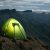 A glowing lime-green tent secured with small rocks on a rocky cliff ledge, overlooking steep, serrated mountain ridges and a sea of low clouds beneath a dim, cloud-streaked sky.