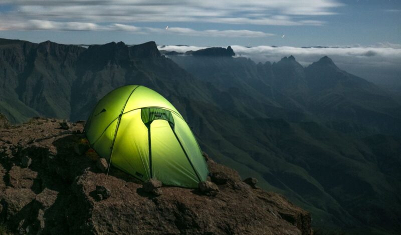 A glowing lime-green tent secured with small rocks on a rocky cliff ledge, overlooking steep, serrated mountain ridges and a sea of low clouds beneath a dim, cloud-streaked sky.