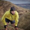 Runner wearing a bright yellow jacket, black beanie, gloves and shorts leans forward with hands on his knees, breathing heavily while pausing on a windswept grassy hillside with rolling hills in the background.