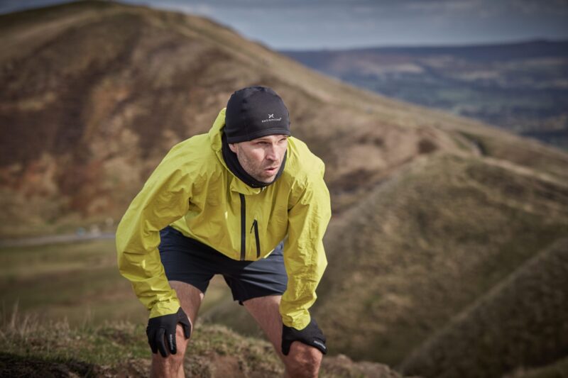 Runner wearing a bright yellow jacket, black beanie, gloves and shorts leans forward with hands on his knees, breathing heavily while pausing on a windswept grassy hillside with rolling hills in the background.