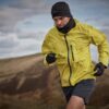 Man mid-stride running uphill in a bright yellow windproof jacket with a black beanie, neck gaiter and gloves across windswept brown moorland under a cloudy sky.
