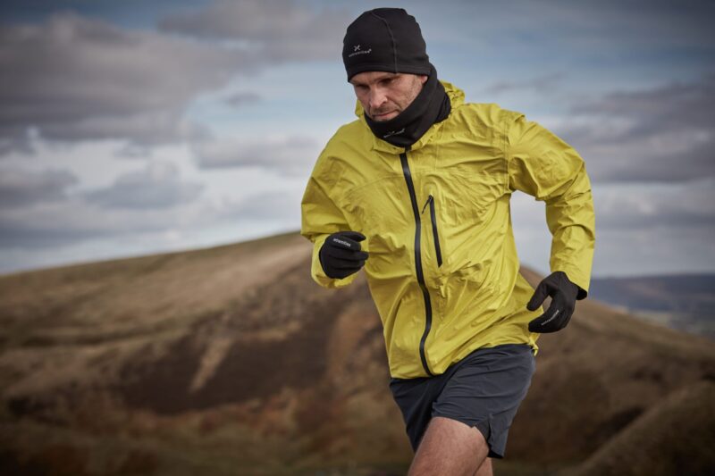 Man mid-stride running uphill in a bright yellow windproof jacket with a black beanie, neck gaiter and gloves across windswept brown moorland under a cloudy sky.