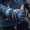 Close-up of gloved hands wearing grey-blue gloves with black leather panels and orange stitching gripping cork-handled trekking poles against a blurred outdoor background.