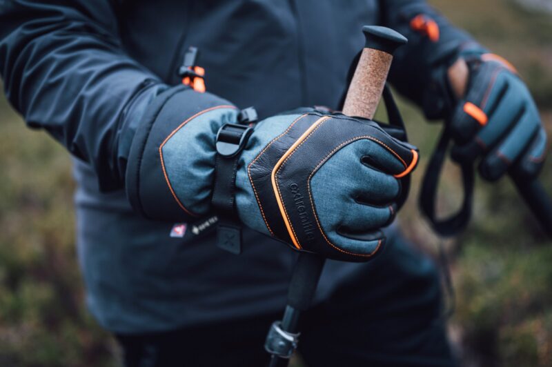 Close-up of gloved hands wearing grey-blue gloves with black leather panels and orange stitching gripping cork-handled trekking poles against a blurred outdoor background.