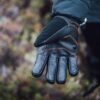 Close-up of an outstretched black insulated glove with leather palm and orange stitching on a person's hand against a blurred natural background.