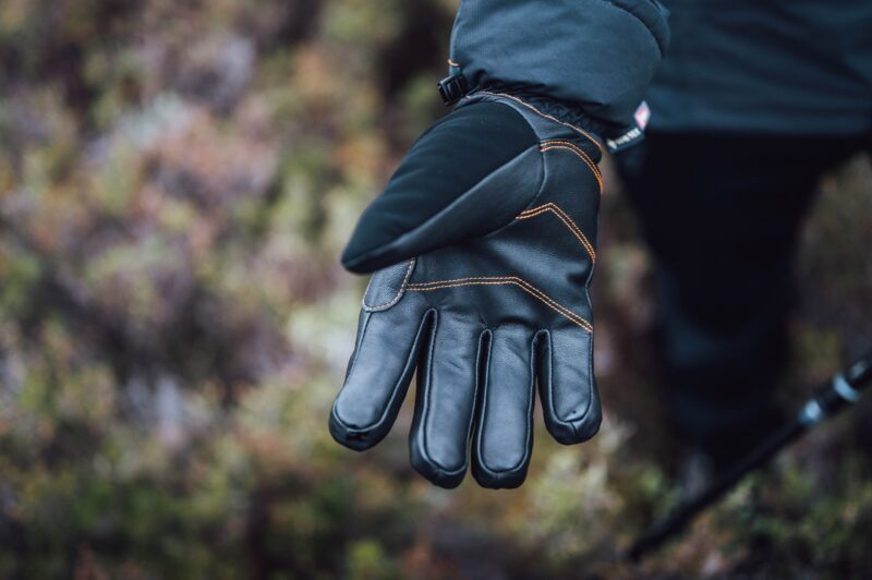 Close-up of an outstretched black insulated glove with leather palm and orange stitching on a person's hand against a blurred natural background.