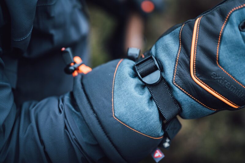 Close-up of a blue and black insulated winter mitten with orange contrast stitching and a black nylon strap and plastic buckle against a blurred outdoor background.