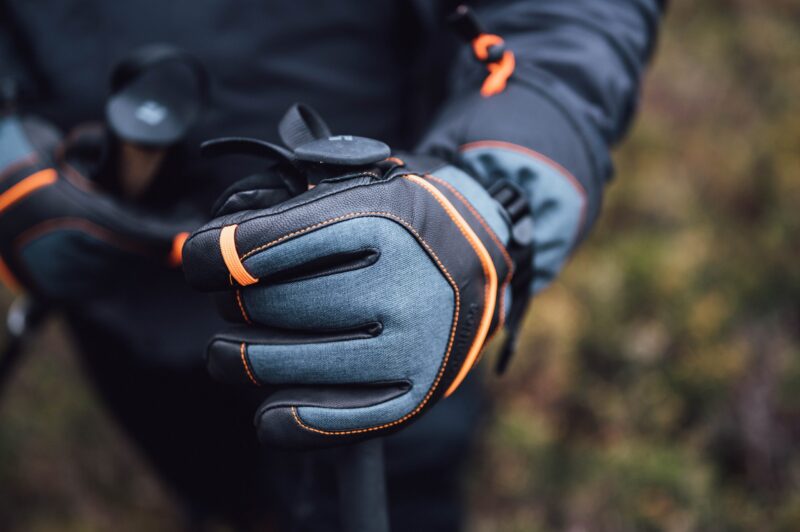Close-up of a grey and black hiking glove with orange stitching and a wrist strap gripping a trekking pole against a blurred outdoor background.