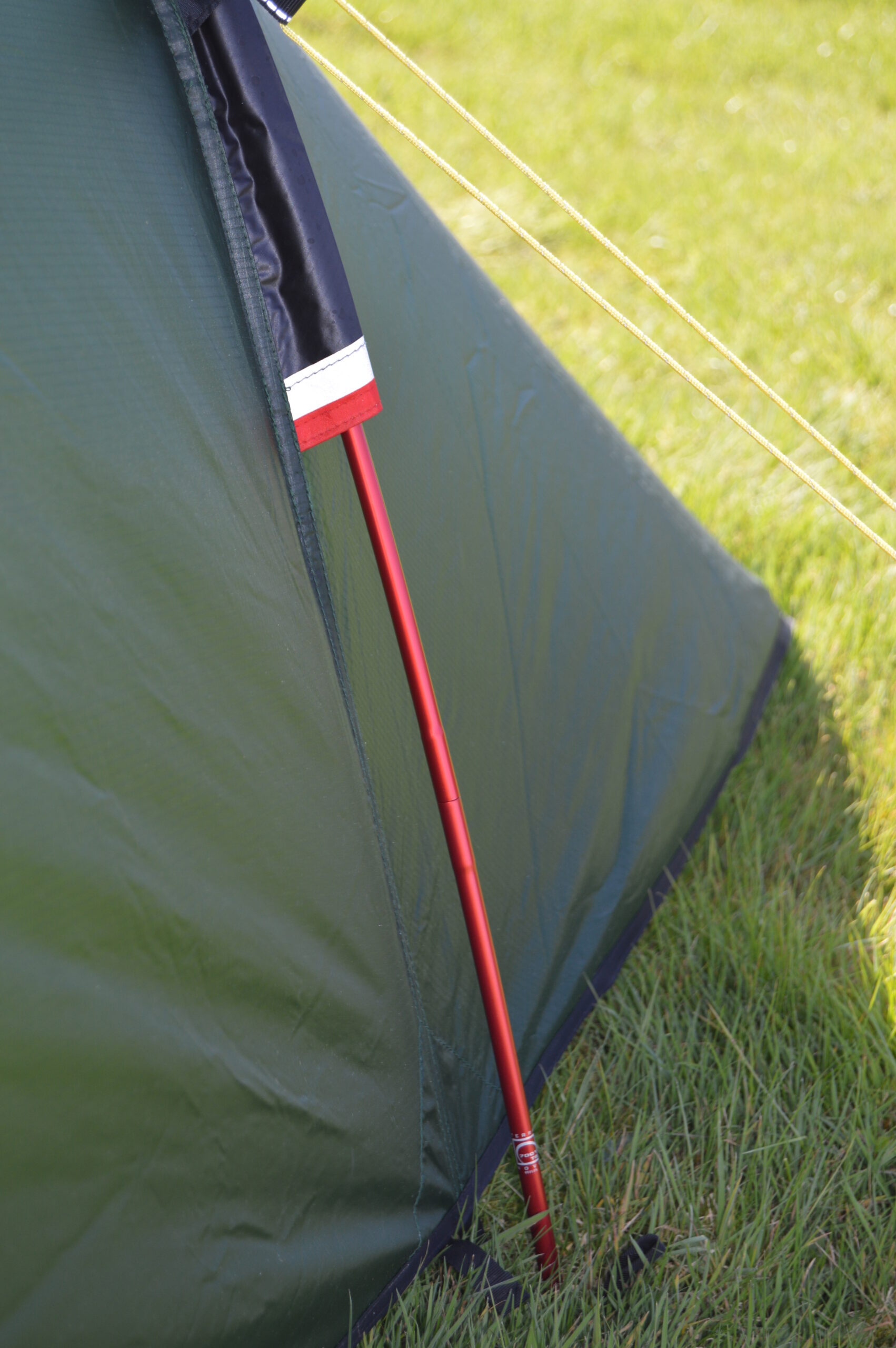 Close-up of a green tent side with a red aluminium pole threaded through a fabric sleeve, two yellow guy lines stretched across sunlit grass and the tent hem pegged into the ground