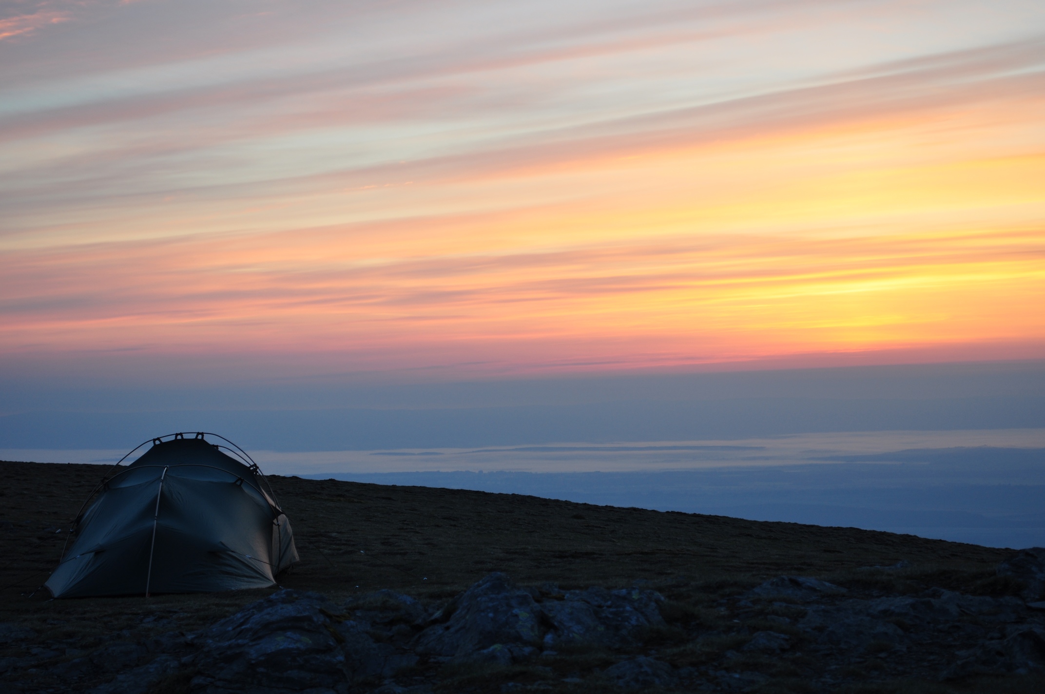 A lone green tent pitched on a rocky hillside beneath a wide pastel-coloured sunset sky streaked with orange, pink and purple above distant lowland layers.