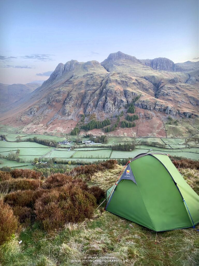 Green camping tent set against a stunning mountainous landscape with rolling hills and valleys under a clear sky, ideal for outdoor enthusiasts and nature lovers.
