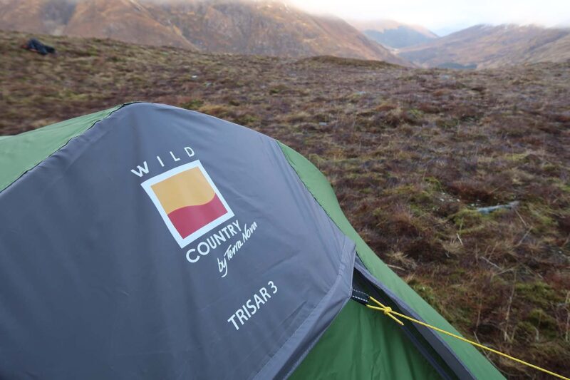 Green three-person tent with a grey rainfly showing a Wild Country by Terra Nova TRISAR 3 logo, pitched on mossy upland with rolling hills in the distance.