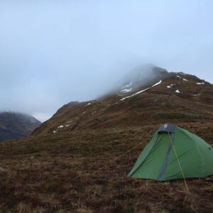 A small green backpacking tent staked on a brown grassy hillside with trekking poles beside it, set against mist‑shrouded, snow‑patched mountain slopes under an overcast sky.
