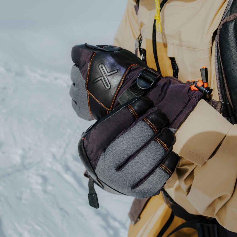 Close-up of insulated grey and black winter gloves with orange stitching and an embossed X logo being adjusted over a tan jacket sleeve against a snowy background.