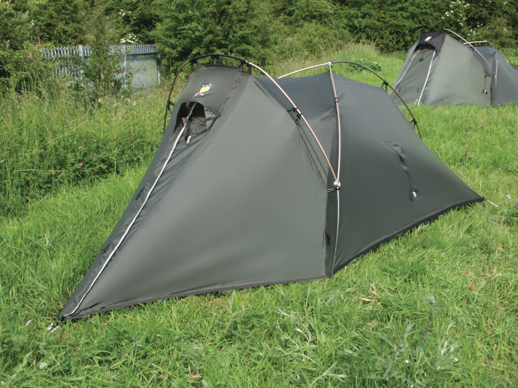 Low-profile olive-green two-person tunnel tent with crossed arched poles and a zippered triangular entrance pitched on a grassy field, another similar tent and bushes visible in the background.