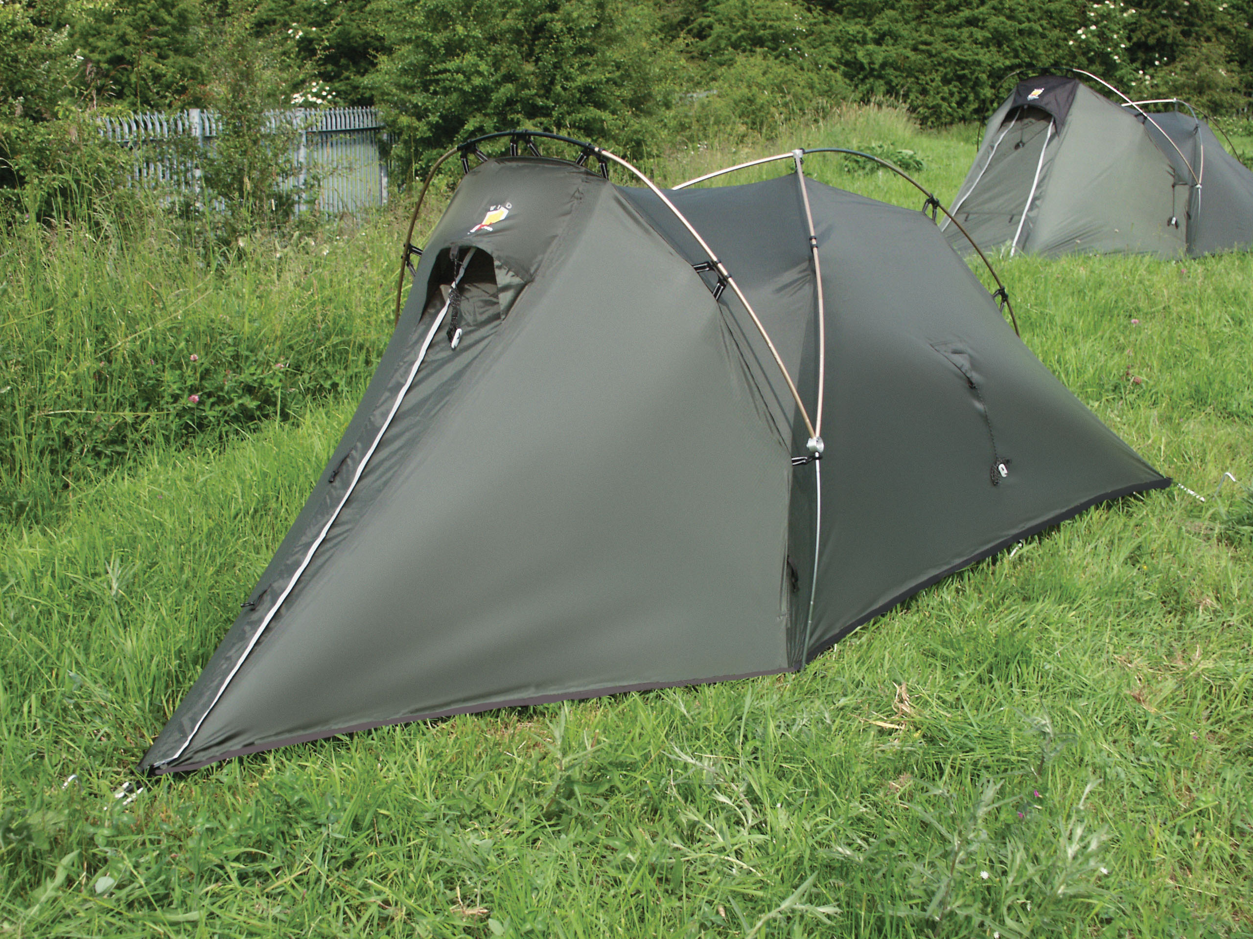 Low-profile olive-green two-person tunnel tent with crossed arched poles and a zippered triangular entrance pitched on a grassy field, another similar tent and bushes visible in the background.
