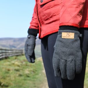 Close-up of a person wearing a red quilted jacket and grey knitted gloves with a tan "extremities" label, standing on a grassy country path beside a wooden fence and distant hills under a blue sky.