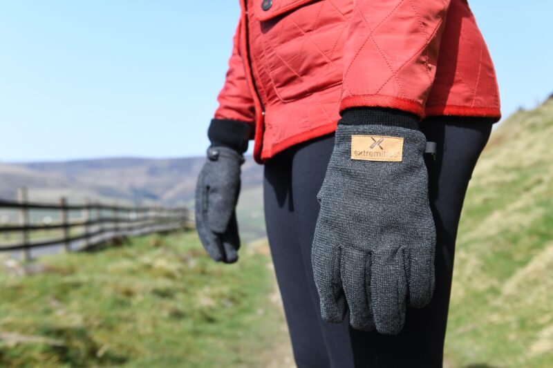 Close-up of a person wearing a red quilted jacket and grey knitted gloves with a tan "extremities" label, standing on a grassy country path beside a wooden fence and distant hills under a blue sky.