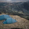 Two teal camping tents pitched on a windswept grassy ridge overlooking steep, rocky mountains and valleys under a muted overcast sky.
