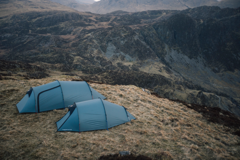 Two teal camping tents pitched on a windswept grassy ridge overlooking steep, rocky mountains and valleys under a muted overcast sky.