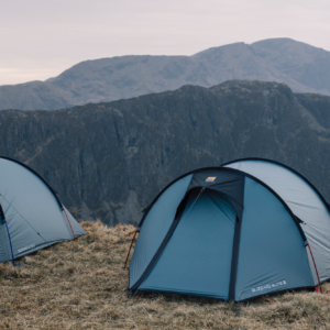 Two blue dome tents pitched on a windswept grassy ridge with steep grey mountain cliffs and an overcast sky in the background.