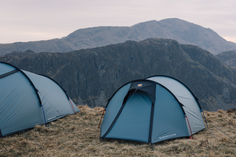 Two blue dome tents pitched on a windswept grassy ridge with steep grey mountain cliffs and an overcast sky in the background.