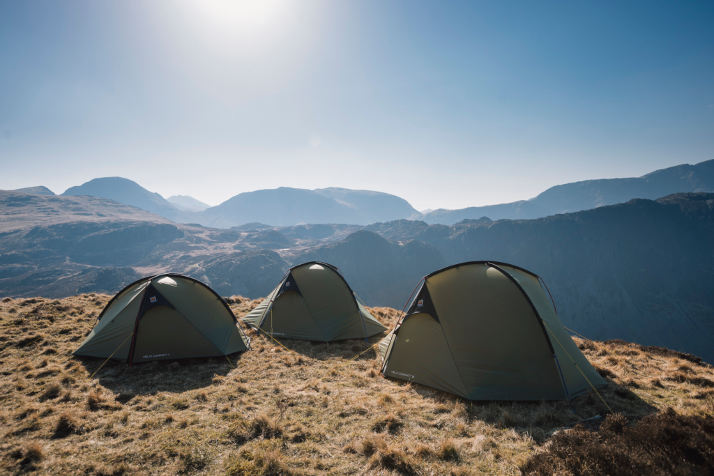 Three olive-green dome tents pitched on a sunlit grassy ridge overlooking layered blue-grey mountains beneath a clear pale-blue sky.