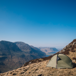 A green two-person tent pitched on a grassy mountain slope beside rocky terrain, overlooking a wide valley of rugged peaks and a distant lake beneath a clear blue sky.