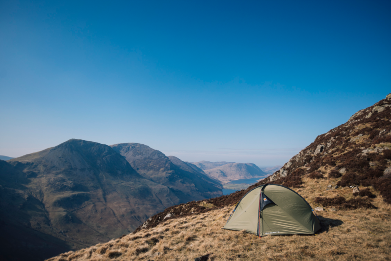 A green two-person tent pitched on a grassy mountain slope beside rocky terrain, overlooking a wide valley of rugged peaks and a distant lake beneath a clear blue sky.