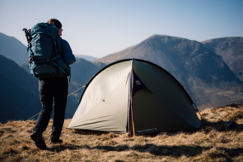 Hiker carrying a large blue backpack stands beside a khaki-green dome tent pitched on a grassy mountain ridge, with sunlit rugged peaks and a clear pale-blue sky in the background.