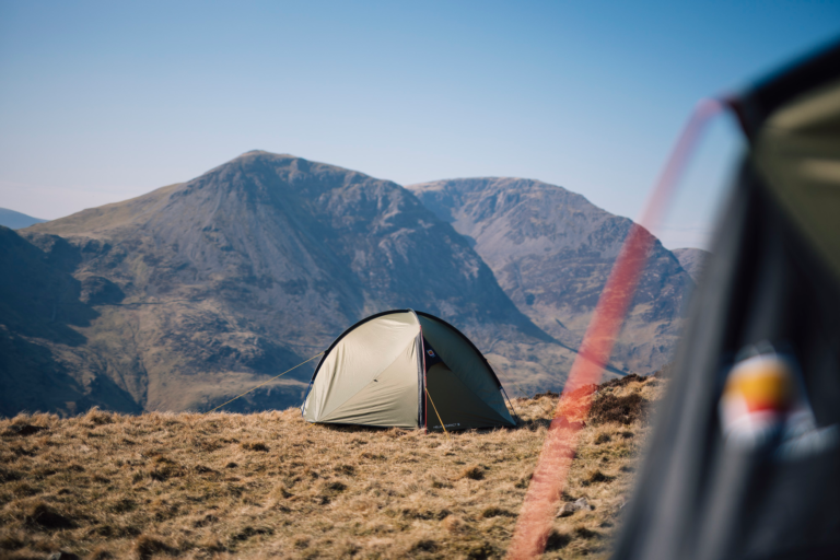 A small green dome tent pitched on a windswept grassy ridge with steep rocky mountains under a clear blue sky, a blurred tent edge and red guyline visible in the foreground.