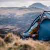 Camper in a blue HELM ELITE 1 tent sits at the open entrance wearing a mustard jacket and handling a small tool, surrounded by grassy tussocks with hazy mountains in the background.
