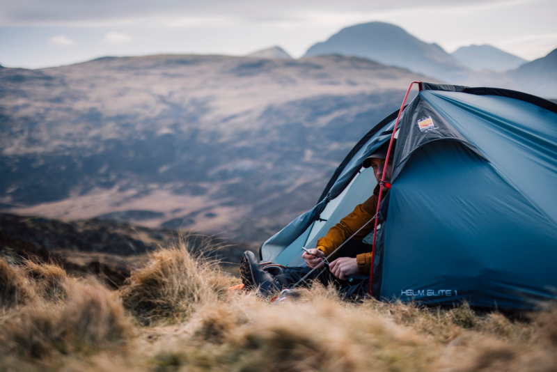 Camper in a blue HELM ELITE 1 tent sits at the open entrance wearing a mustard jacket and handling a small tool, surrounded by grassy tussocks with hazy mountains in the background.