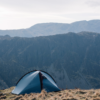 Blue two-person tent pitched on a grassy ridge near a cliff edge with steep, layered mountain cliffs and a pale sky in the background.