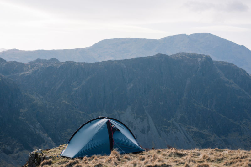 Blue two-person tent pitched on a grassy ridge near a cliff edge with steep, layered mountain cliffs and a pale sky in the background.
