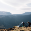 A camper crouches beside a small grey tent pitched on a windswept grassy ridge overlooking layered blue-grey mountains and a pale sky.