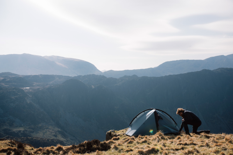 A camper crouches beside a small grey tent pitched on a windswept grassy ridge overlooking layered blue-grey mountains and a pale sky.