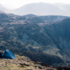 A lone camper crouches beside a small blue tent on windswept grassy slopes with steep rocky cliffs and hazy, layered mountains rising in the distance.