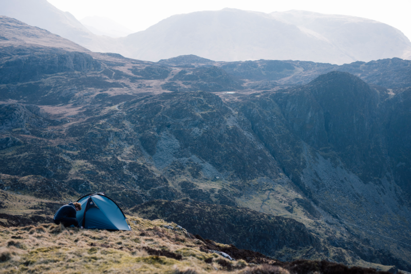A lone camper crouches beside a small blue tent on windswept grassy slopes with steep rocky cliffs and hazy, layered mountains rising in the distance.