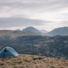 Small blue dome tent pitched on a tussock-covered hillside, with layered rocky hills and distant misty mountains beneath a pale, overcast sky.