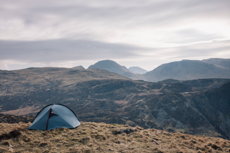 Small blue dome tent pitched on a tussock-covered hillside, with layered rocky hills and distant misty mountains beneath a pale, overcast sky.