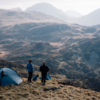 Two people and a dog stand beside blue tents on a grassy mountain ridge, looking out over layered rocky hills and misty peaks under a pale sky.